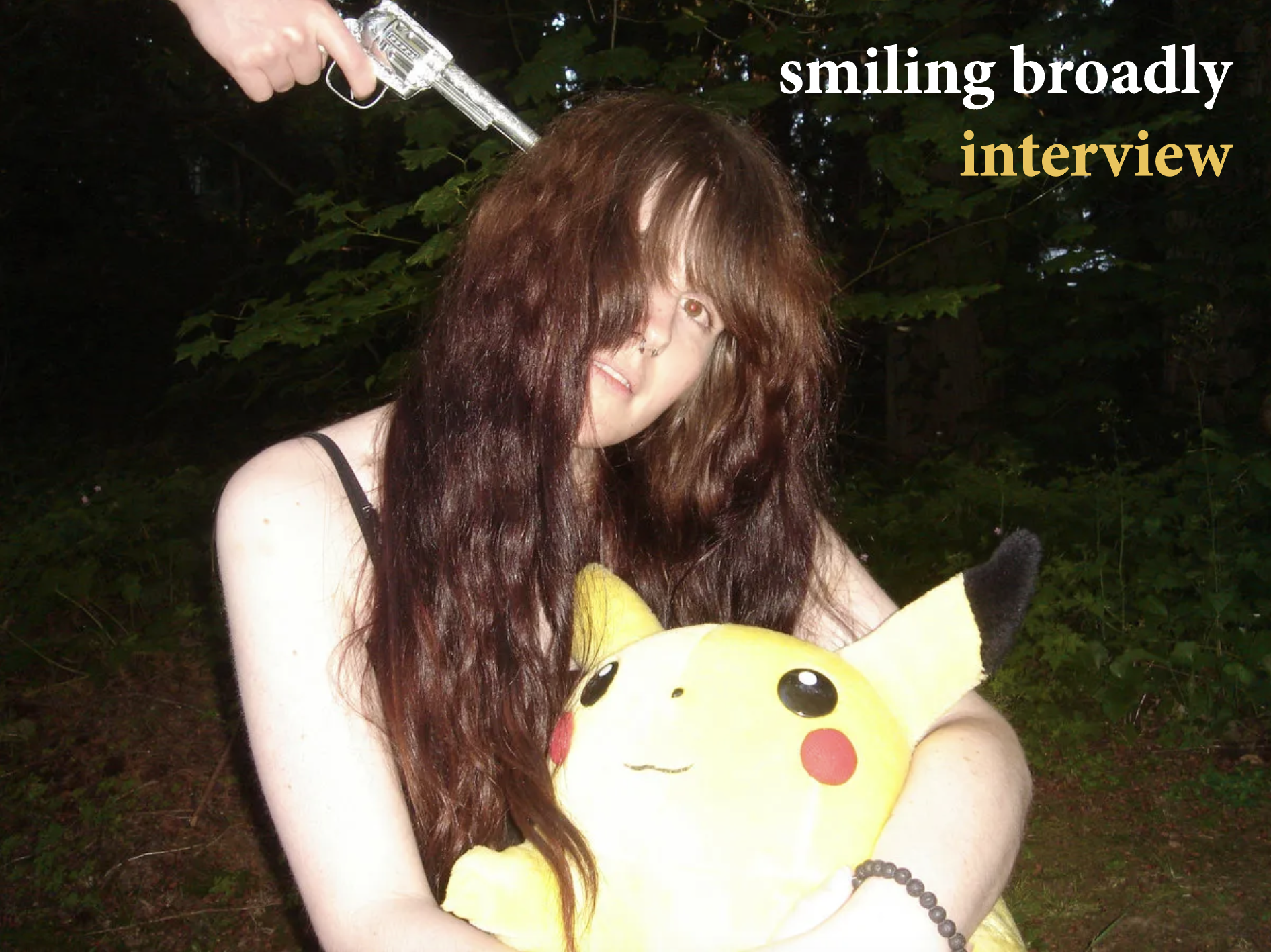 Hazel of smiling broadly sits, holding a Pikachu plush. Someone is pointing a gun to her head from just off-screen.