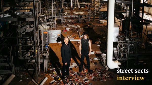 Leo Ashline and Shaun Ringsmuth of Street Sects stand in an abandoned warehouse, with several objects strewn about. The camera is craned high above them, and both figures cast long shadows.